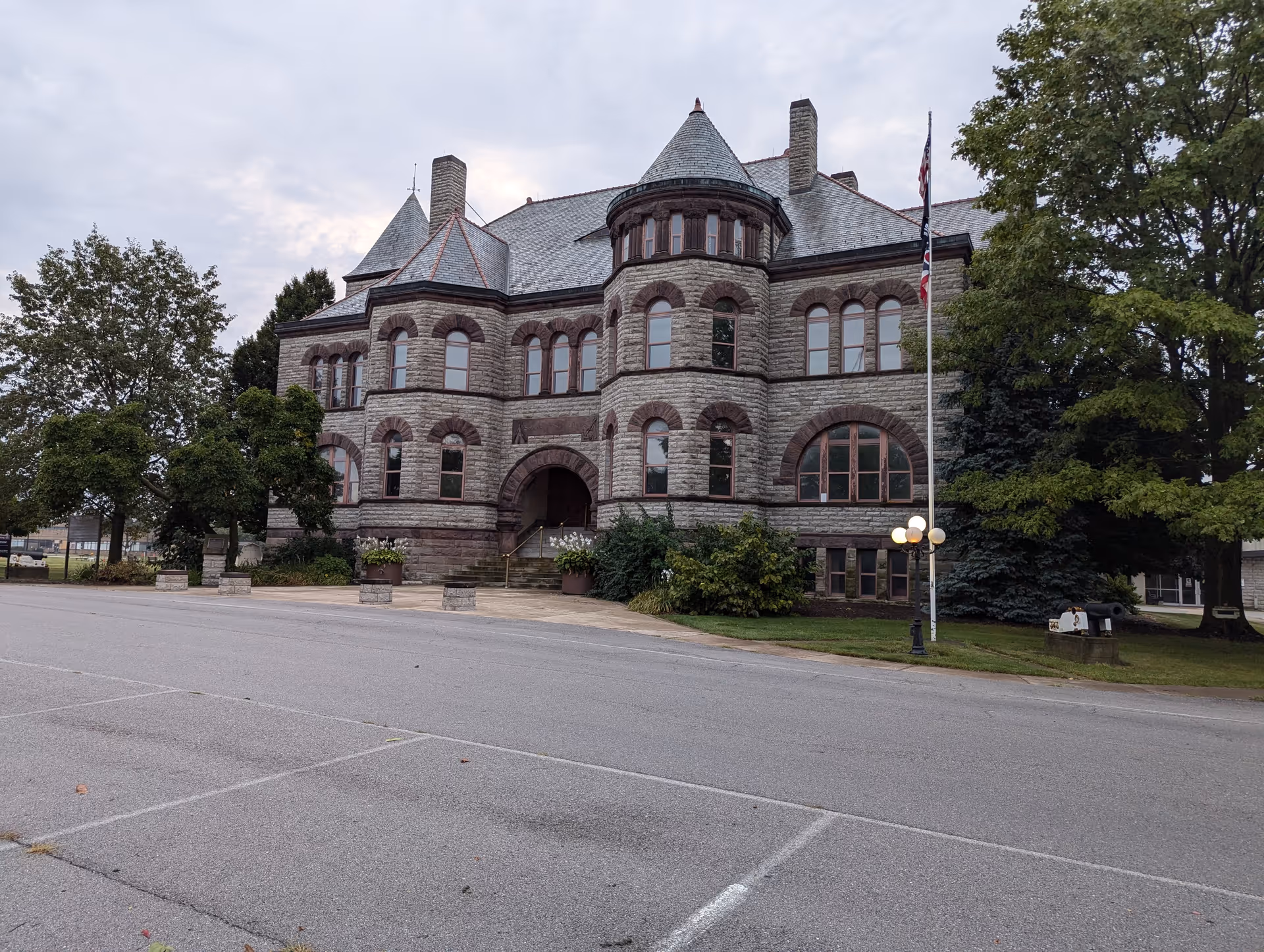 Historic stone building with turrets and a flagpole along a tree-lined street.