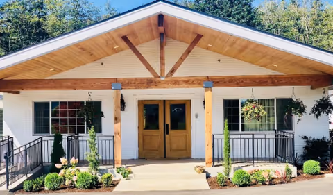 Front exterior view of a single-story building with a peaked roof, wooden double doors, white brick walls, and hanging flower baskets on either side of the entrance. There are small shrubs and plants along the walkway leading to the door.