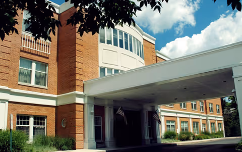 Exterior front entrance of a brick senior living facility with a covered porte-cochère, American flags, windows, and landscaped beds under a blue sky.
