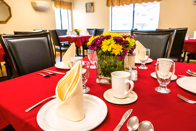 A dining table with a red tablecloth set with plates, glassware, a folded napkin and a vase of yellow and purple flowers in a dining room.