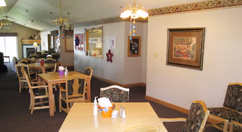 Interior view of a senior living facility dining area with several wooden tables and chairs. The room has carpeted floors, framed artwork on the walls, and decorative hanging lights. In the background, there is a fireplace and additional seating areas.