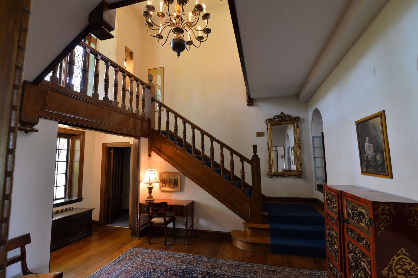 Foyer with a wooden staircase, chandelier, small desk, and ornate mirror in a traditional interior.