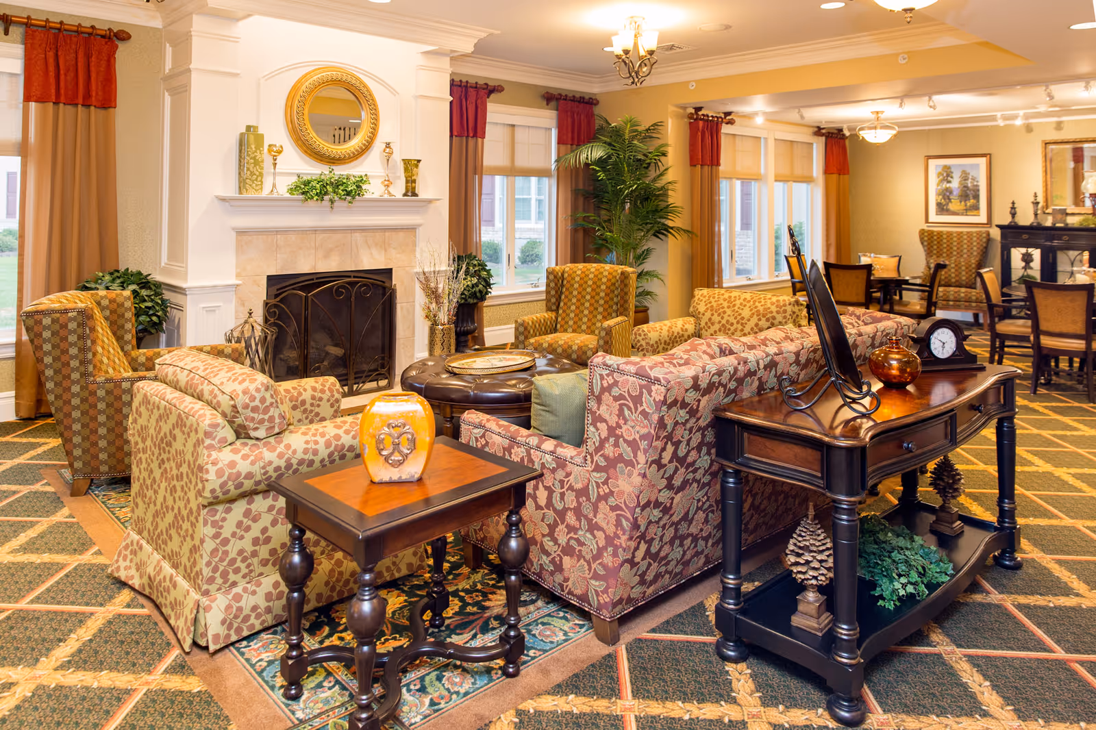 A warm, furnished communal living room with patterned sofas and armchairs arranged around a fireplace and decorative tables.