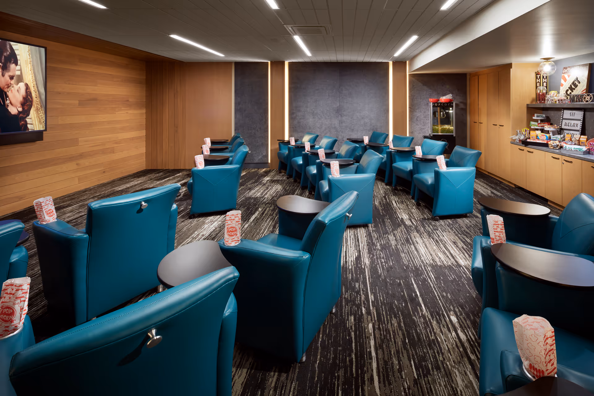 Small screening room with teal recliner seats arranged in rows, side tables and popcorn boxes, a wall-mounted screen and a snack counter.