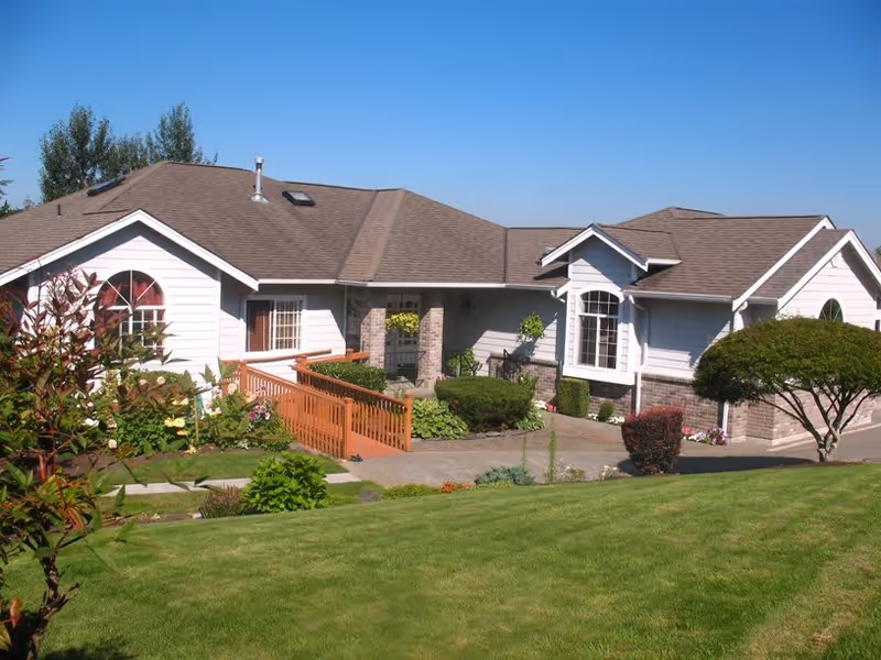 A single-story residential building with white siding and a brown shingled roof, surrounded by a well-maintained lawn and various shrubs and plants. There is a wooden ramp leading to the front entrance, which has a hanging plant. The sky is clear and blue.