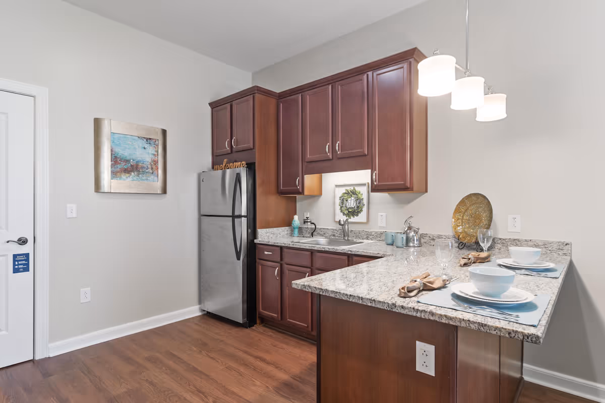 A modern kitchen with dark wooden cabinets, a stainless steel refrigerator, granite countertops, and a small sink. The countertop extends into a breakfast bar set with two place settings including bowls, plates, glasses, and napkins. There is a decorative plate on a stand and a framed artwork on the wall above the sink. The floor is wooden and the walls are painted light gray.