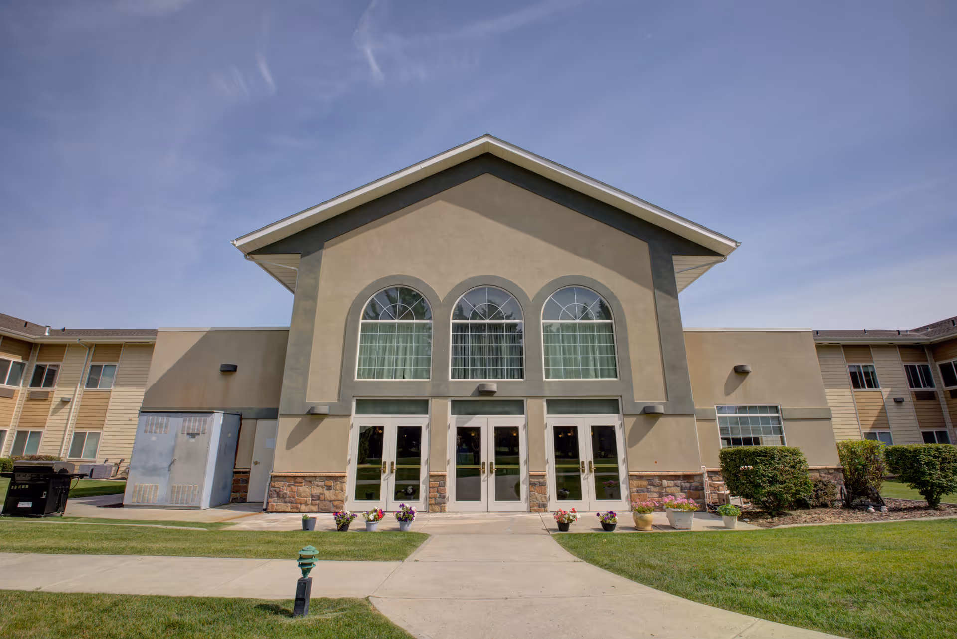 Front exterior view of a senior living facility building with three large arched windows above three sets of double glass doors. The building has beige and stone accents with a well-maintained lawn and flower pots near the entrance under a clear blue sky.