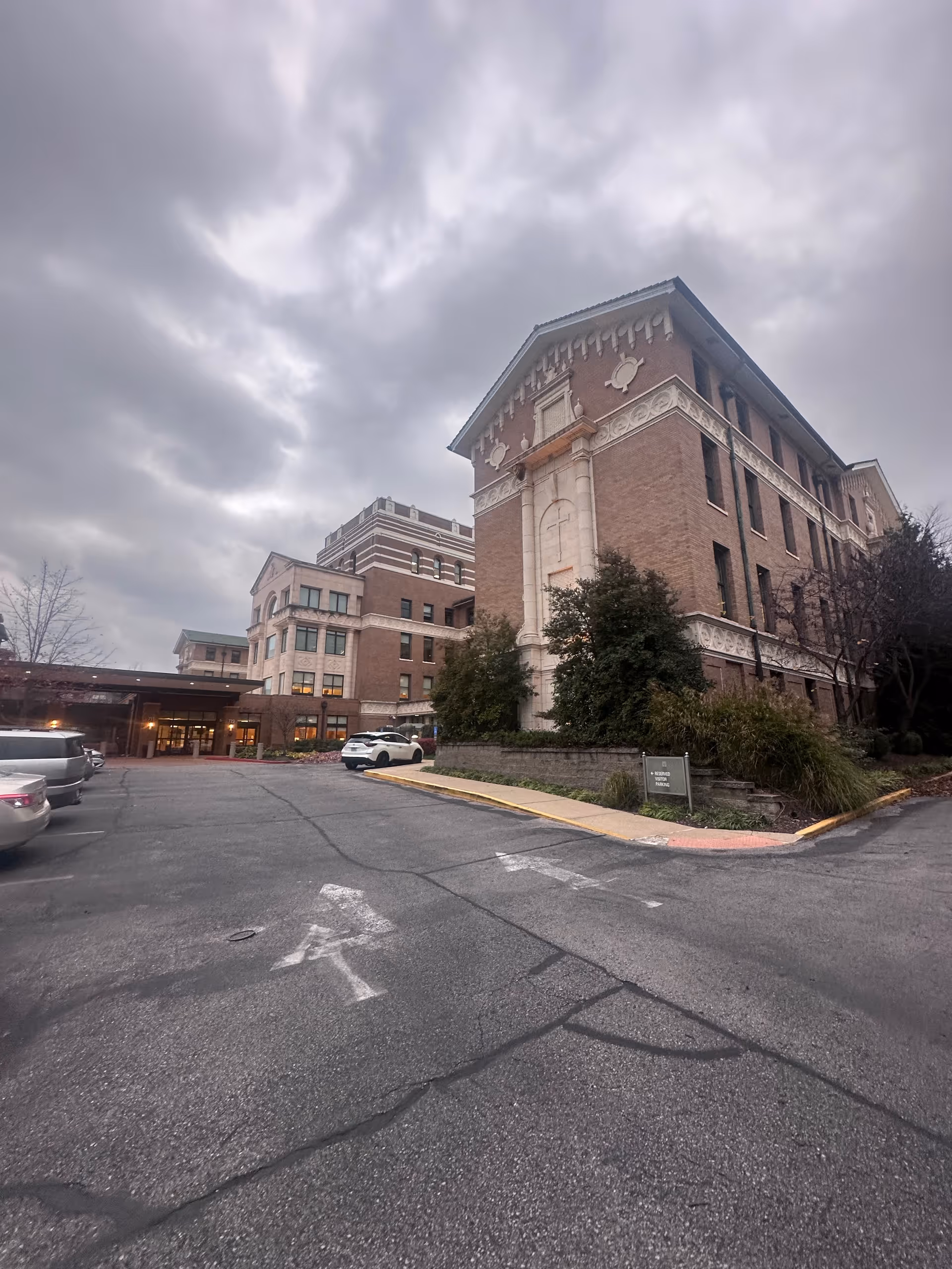Brick multi-story convalescent home building with a driveway, parked cars, and an overcast sky.