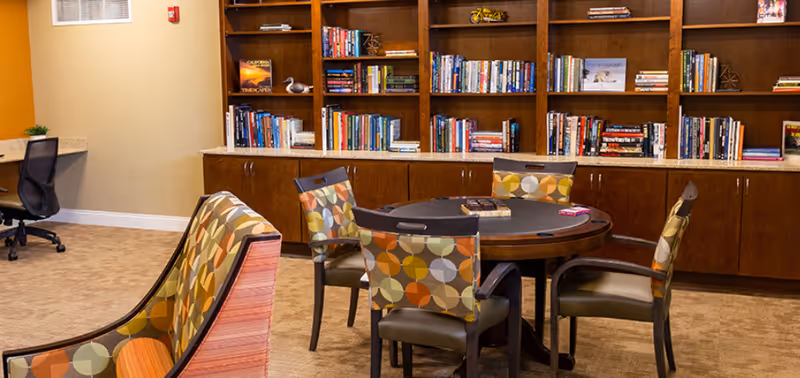 A cozy library or common area with a round table surrounded by four chairs featuring colorful circular patterned upholstery. Behind the table is a large wooden bookshelf filled with books and decorative items. To the left, there is a desk with a black office chair and a small plant on the counter.