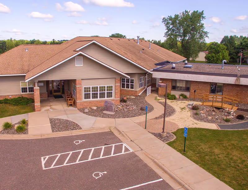 A single-story beige senior living facility with accessible entrance and landscaped front, parking lot visible.