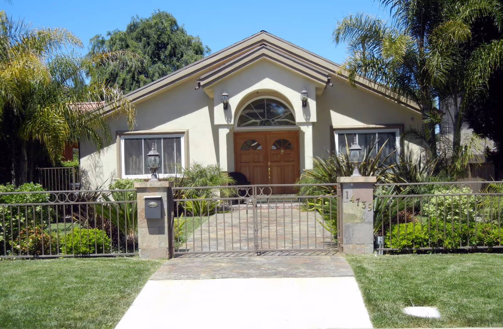 Beige single-story building with arched entry, wooden doors, gated front yard, and palm trees.
