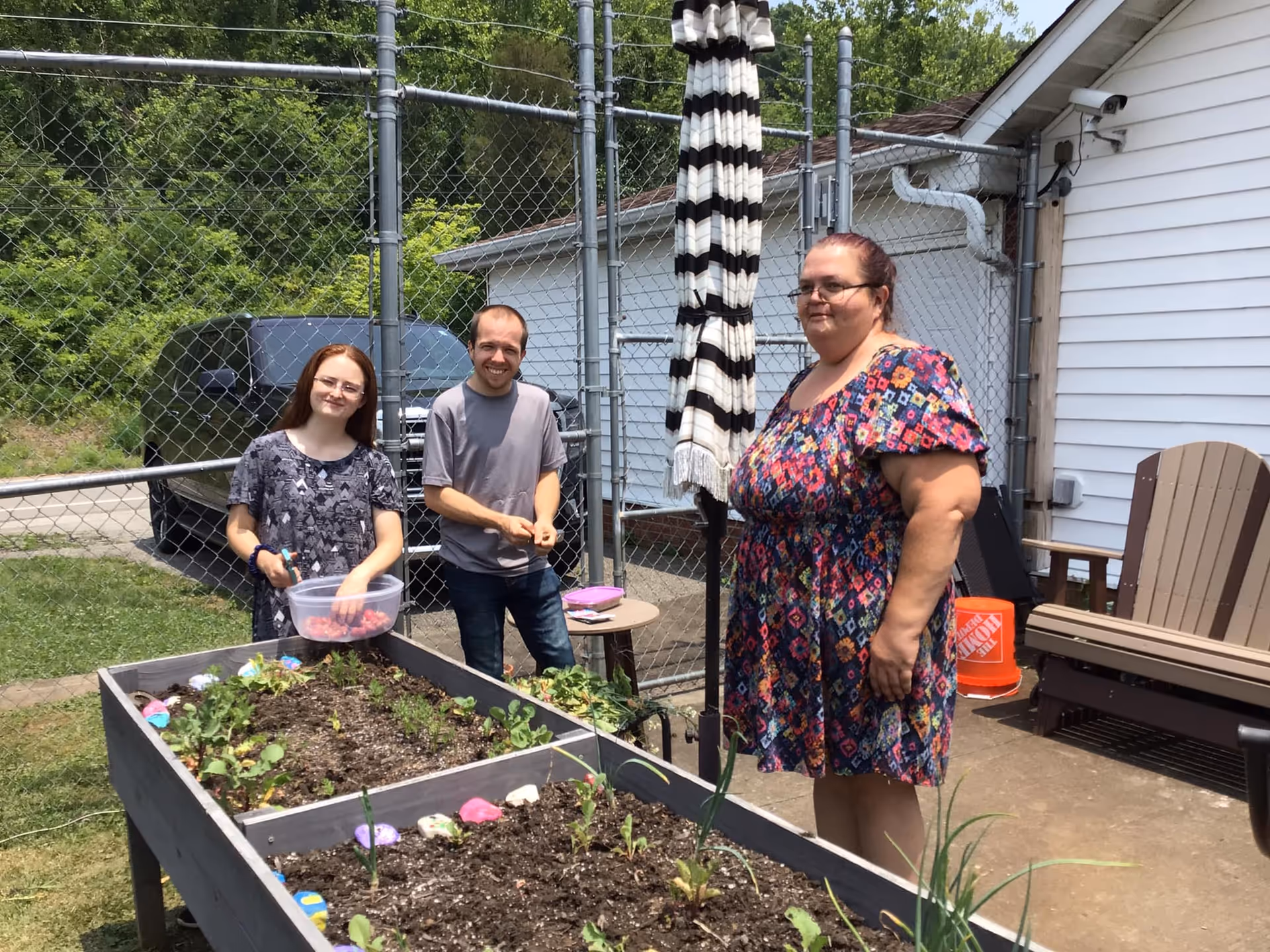 Three people standing outdoors next to a raised garden bed with small plants growing in it. One person is holding a container with red berries, and the other two are standing nearby. Behind them is a chain-link fence, a white building, and outdoor furniture including a striped umbrella and Adirondack chairs.