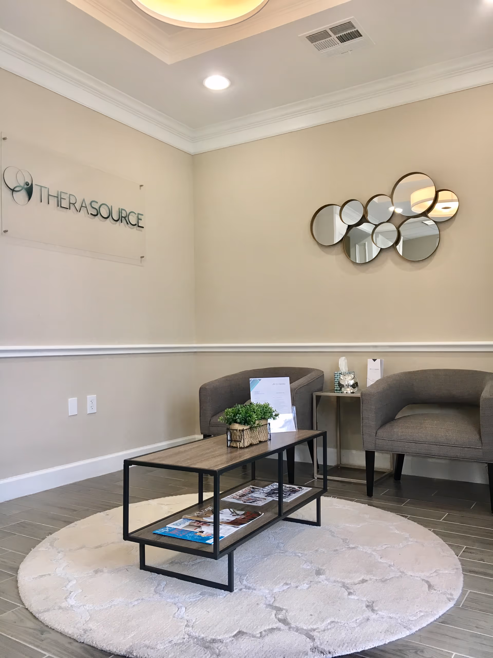 Modern reception lounge with beige walls, circular mirror art, two gray chairs, coffee table, TheraSource sign.