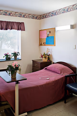 Small single-occupancy bedroom with a single bed dressed in burgundy linens, bedside table and flowers, a window with a valance, and an overbed table.