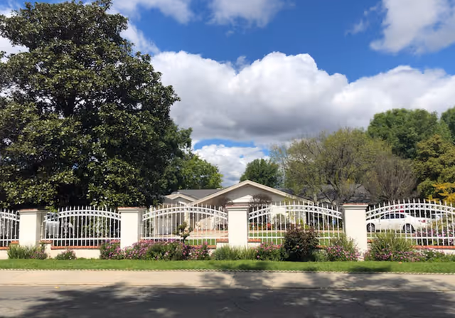 Front view of a gated senior living campus with a white decorative fence and greenery.
