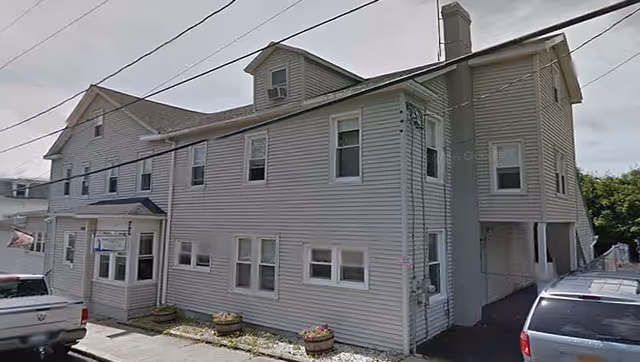 Exterior view of a two-story beige residential building with multiple windows and a small covered entrance. Several vehicles are parked along the street and driveway beside the building. The sky is overcast.