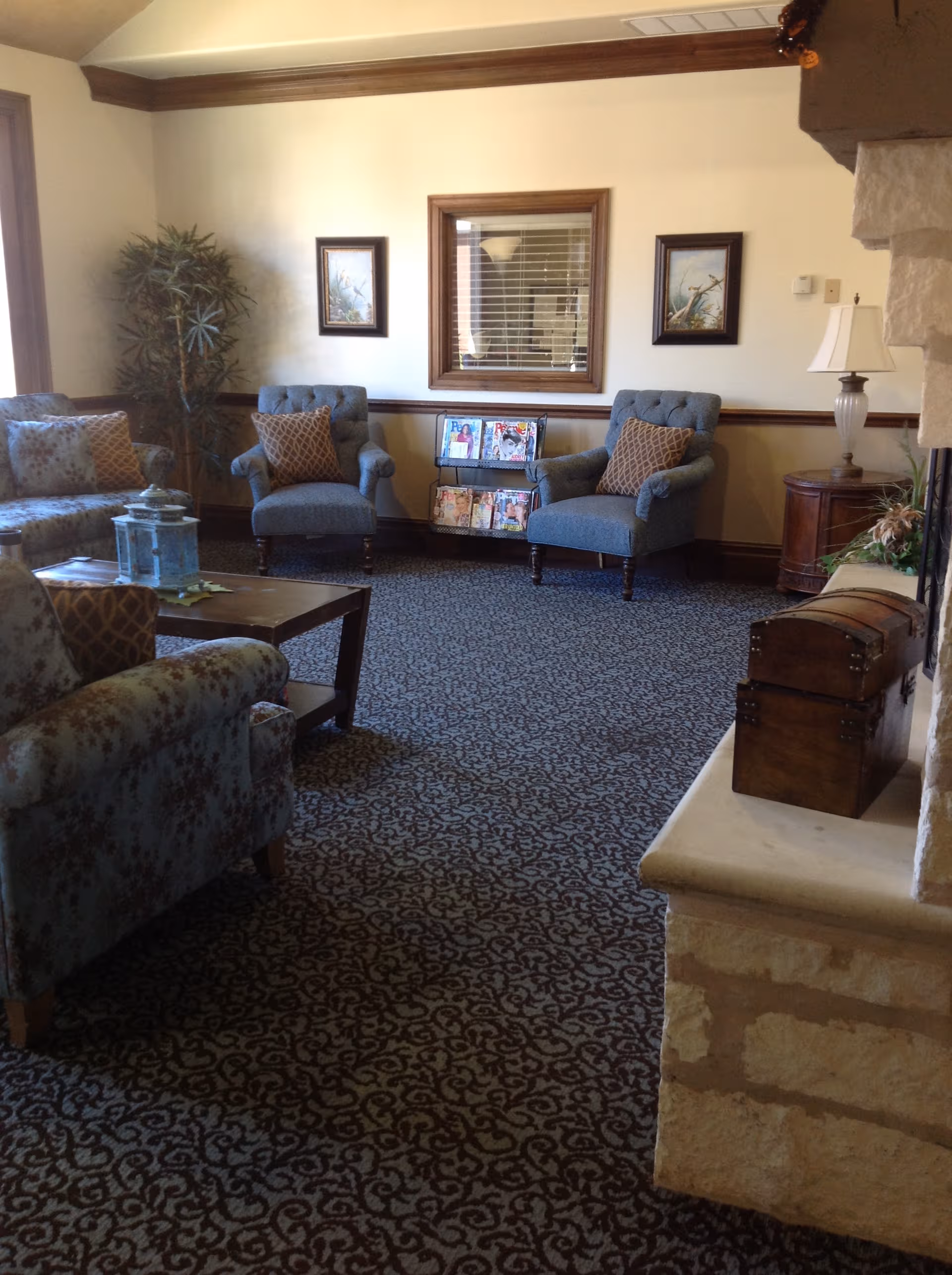 Cozy senior living room with blue chairs, patterned carpet, magazines, and a stone fireplace.