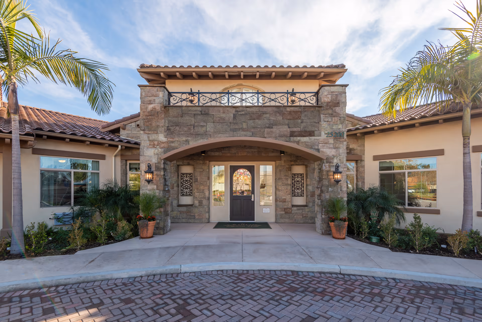 Front entrance of a Mediterranean-style senior living building with a stone facade, arched entryway, potted plants and palm trees.