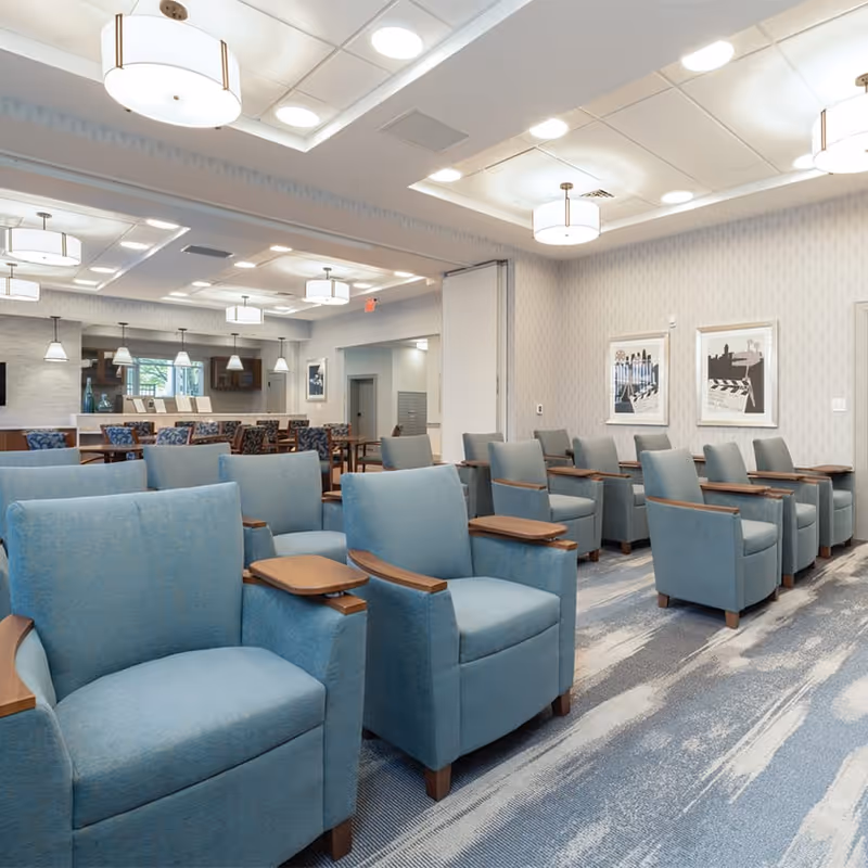 A spacious senior living community room with multiple rows of comfortable blue armchairs featuring wooden armrests and attached small tables. The room has a modern design with patterned carpet, light-colored walls adorned with framed artwork, and ceiling lights providing bright illumination. In the background, there is a dining area with tables and chairs, pendant lights, and a window.