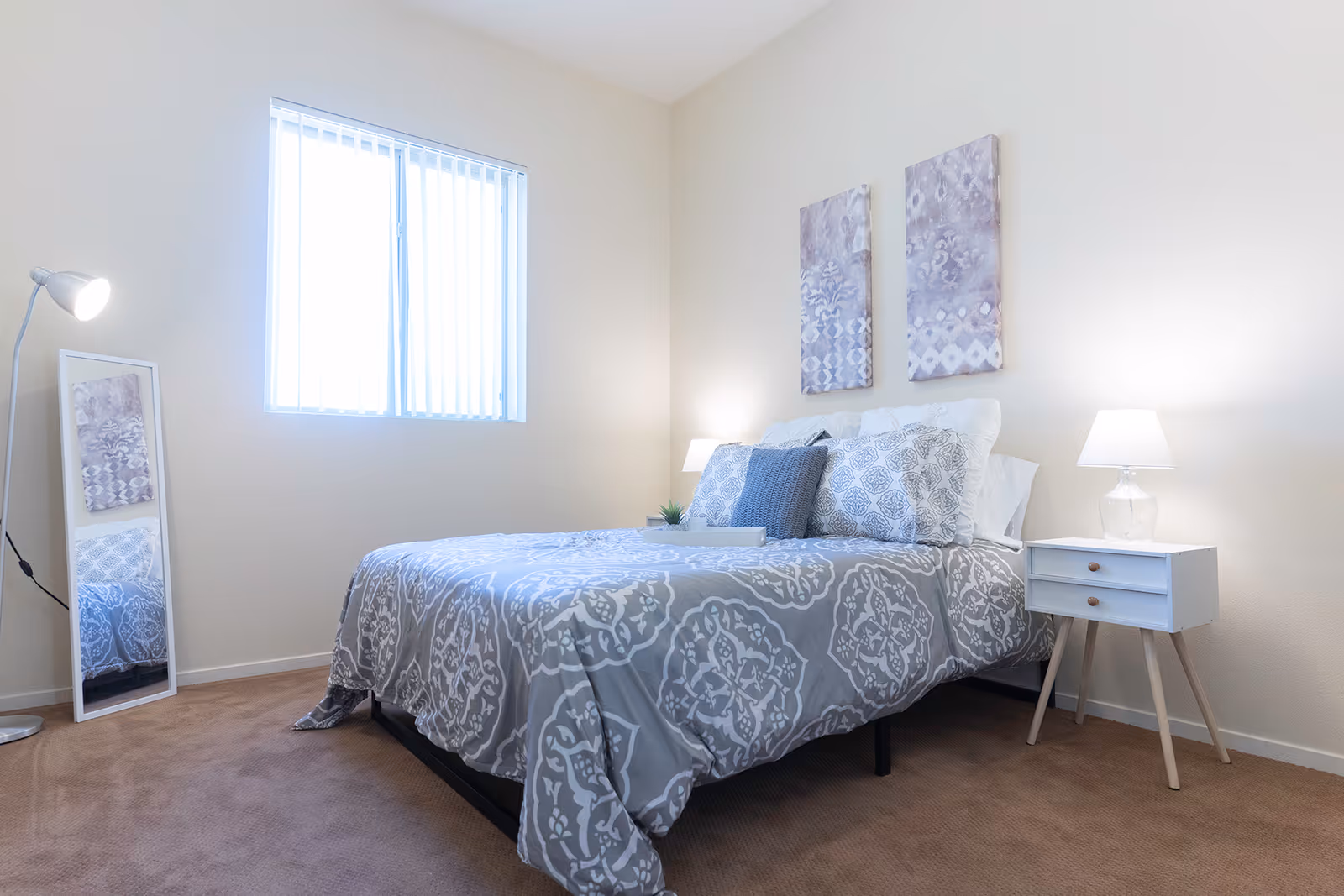 Bright bedroom with a made bed featuring gray patterned bedding, a nightstand and lamp, wall art, and a standing mirror.