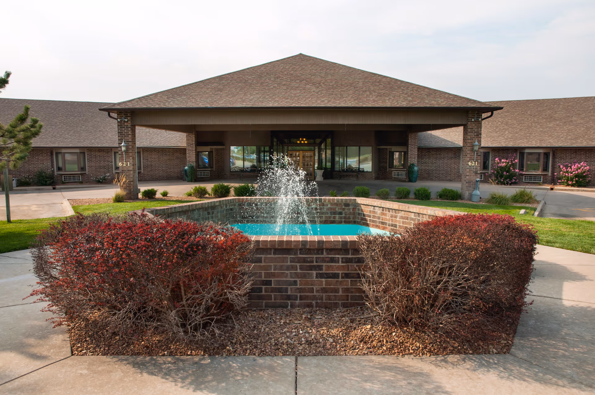 Front exterior view of a single-story brick building with a covered entrance, a small square fountain with water spraying in the center, and landscaped bushes and greenery around the fountain and walkway.