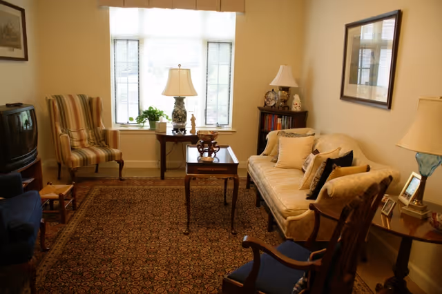 Cozy living room with a sofa, multiple armchairs, a patterned area rug, a central coffee table, lamps, and a large window.