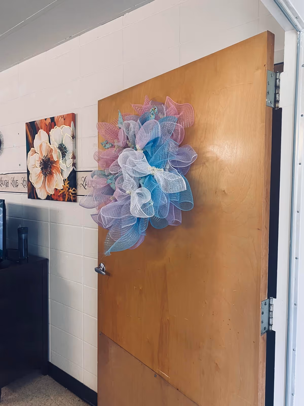 A wooden interior door decorated with a pastel mesh wreath, with floral wall art on a cinderblock wall beside it.
