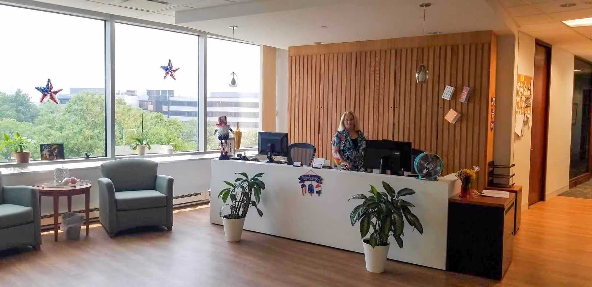 Bright lobby reception with a staff member at a white desk, chairs, plants, and large windows.