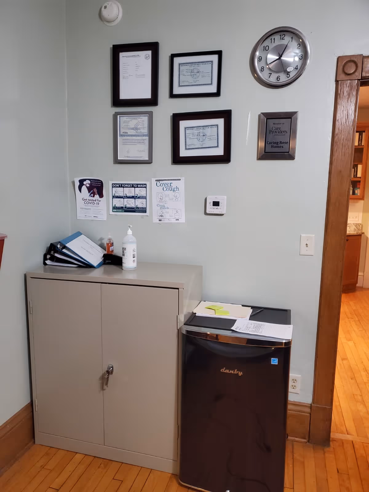 Interior corner with a beige cabinet, black mini-fridge, wall certificates, clock, and posted notices.