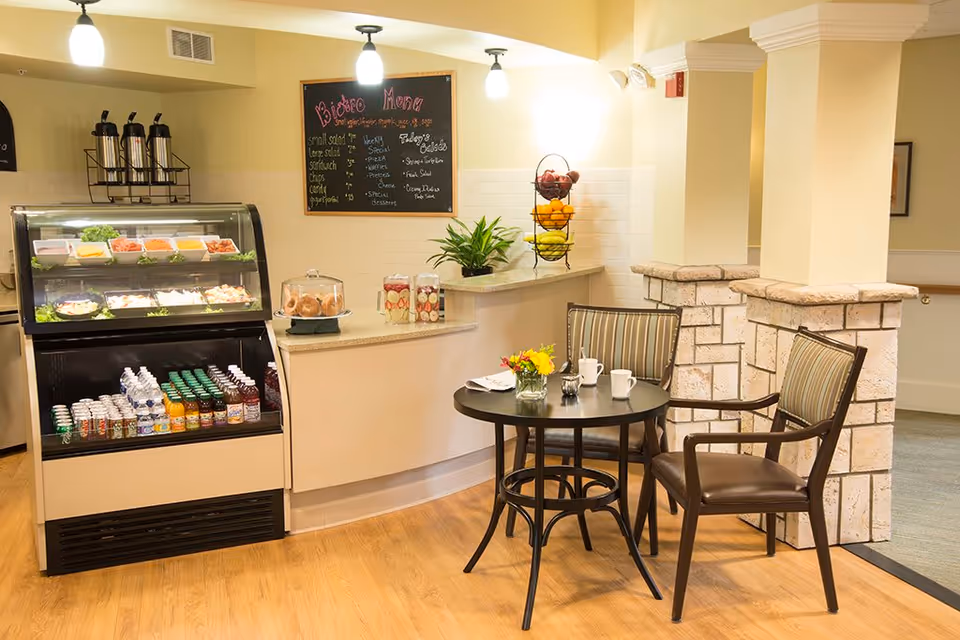 A small dining area in a senior living facility with a round table set with two cups, a small vase of flowers, and two chairs. Behind the table is a counter with a display case containing various salads and drinks. Above the counter is a chalkboard menu listing bistro items and daily specials. The area is warmly lit with hanging lights and decorated with a plant and a tiered fruit basket.