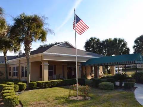 Sunlit senior living facility front with flagpole, palm trees, and a covered entrance.