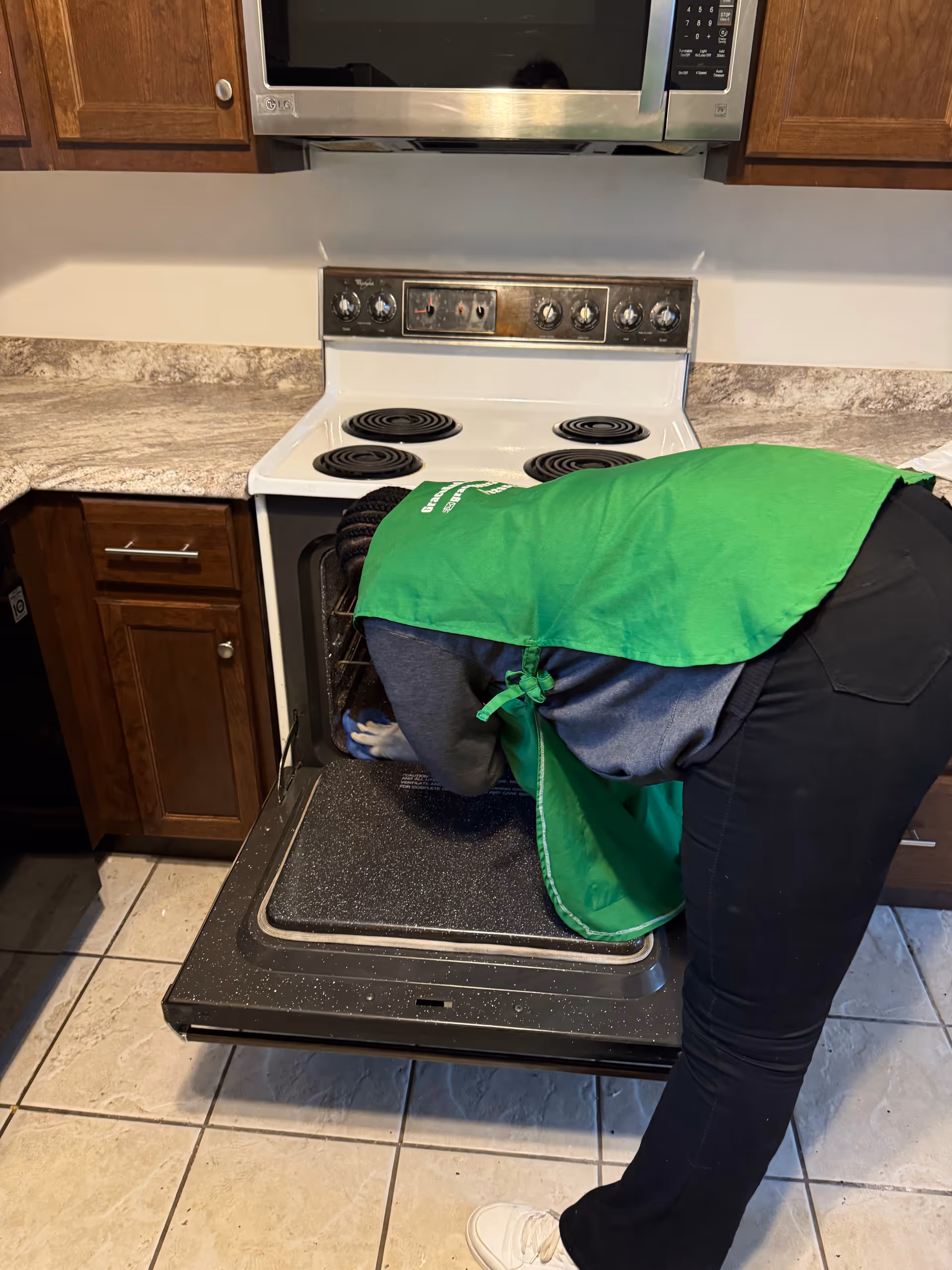 A person in a green apron cleans an oven in a kitchen with wooden cabinets.