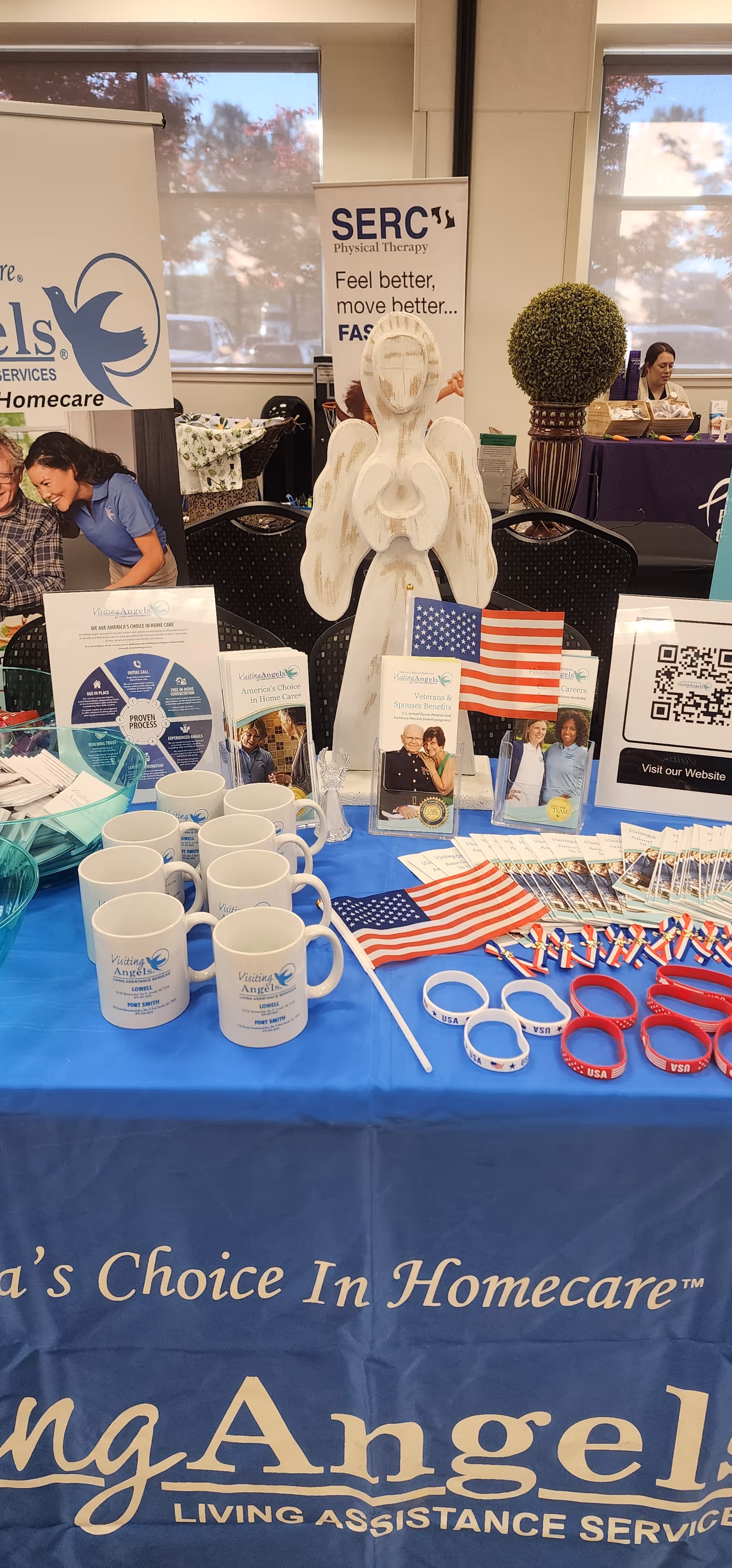 Indoor booth display for Visiting Angels Senior Home Care with banners, mugs, and American flags.