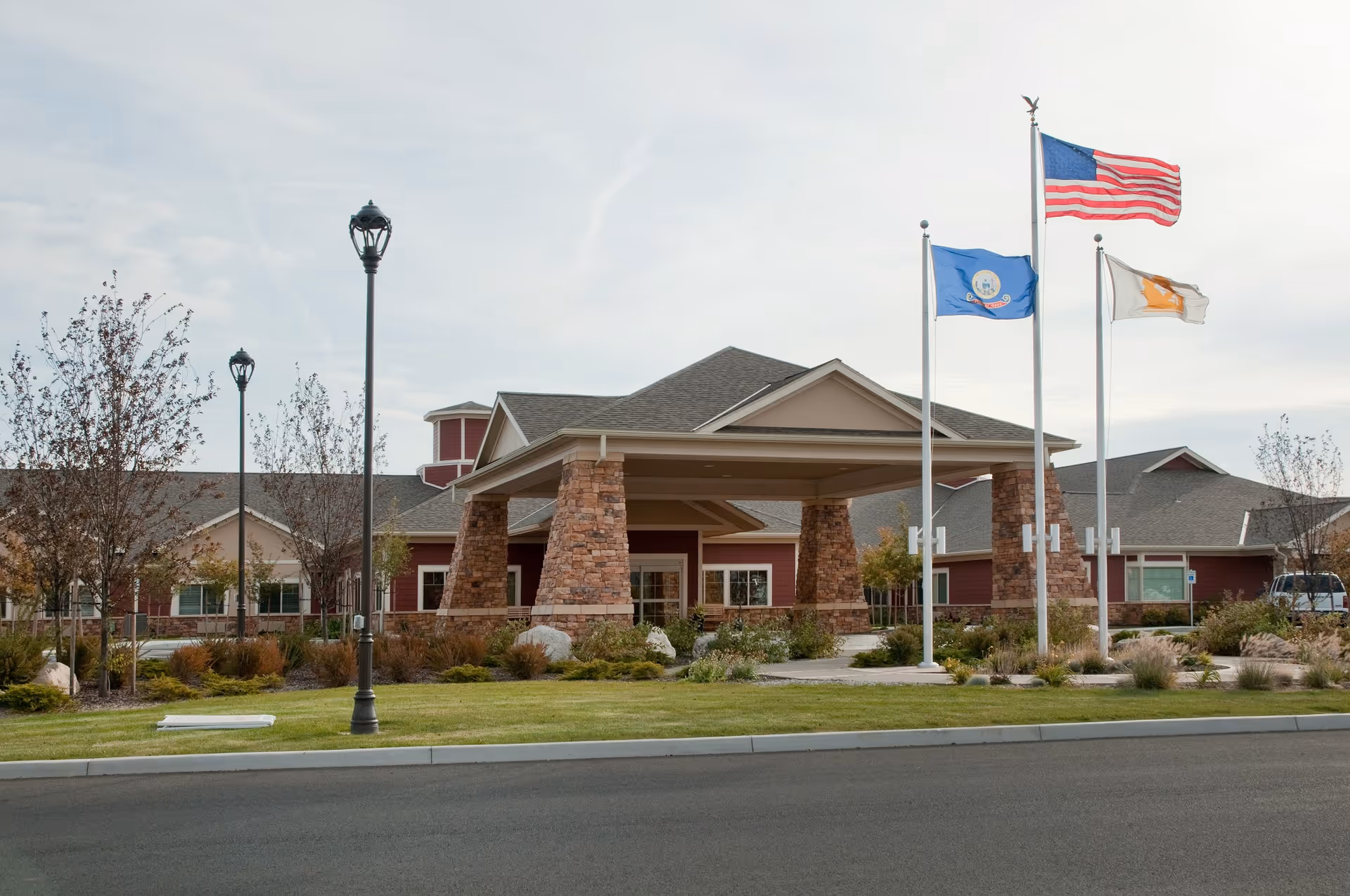 Front exterior view of the Life Care Center of Post Falls building with a covered entrance supported by stone pillars, three flagpoles displaying the American flag, a state flag, and another flag, surrounded by landscaped greenery and a paved road in the foreground.