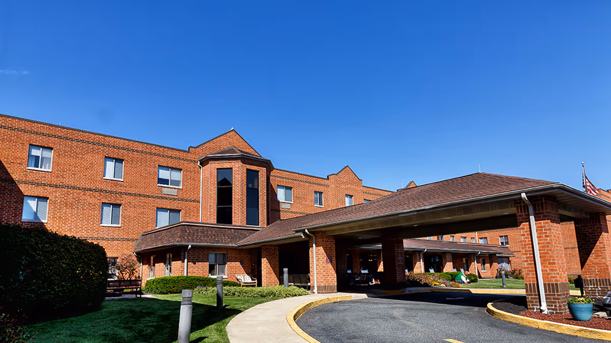 Brick three-story senior living building with a covered drop-off porte-cochère, driveway and manicured lawn under a clear blue sky.