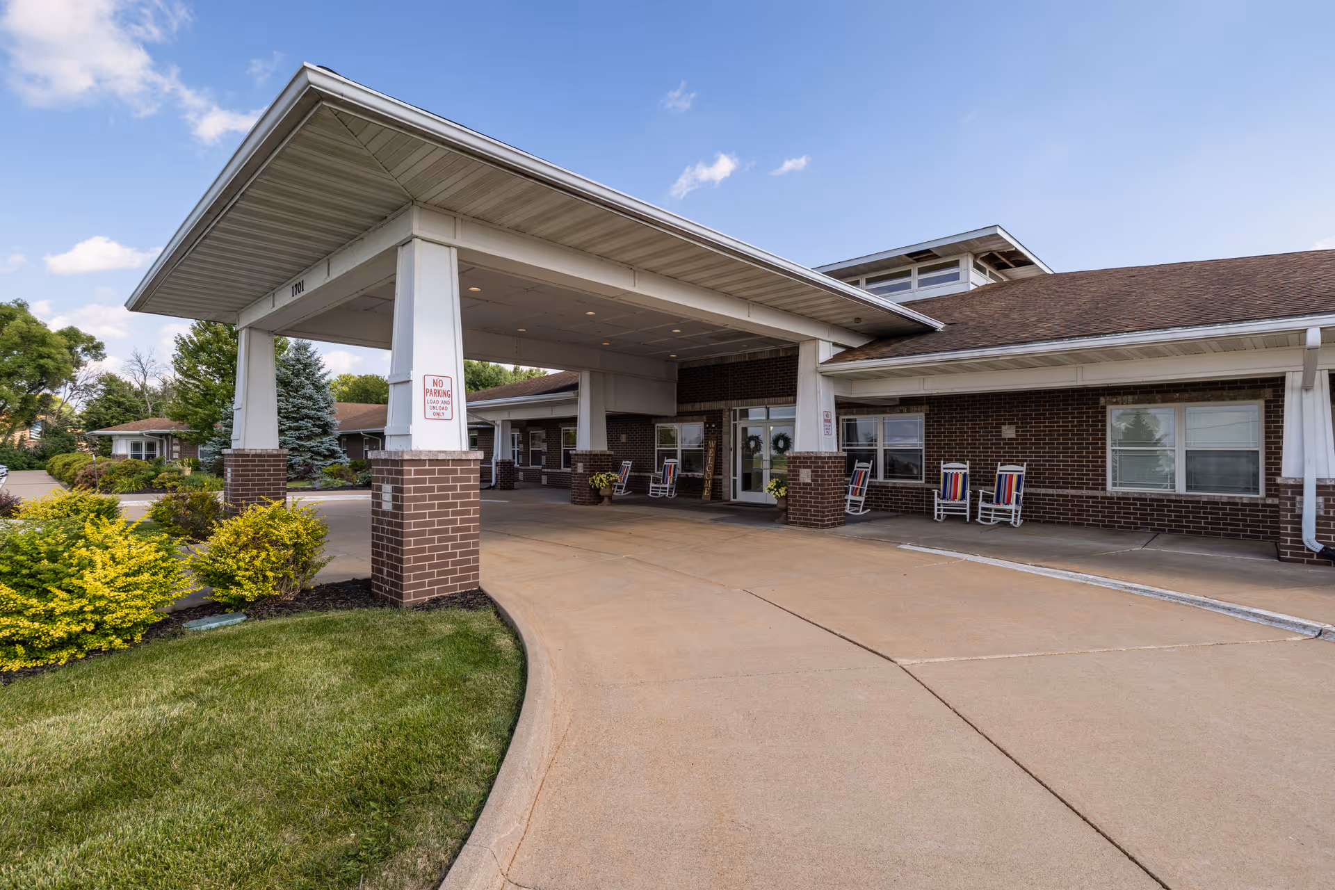 Front entrance and covered driveway of a brick senior living building with rocking chairs and landscaping.