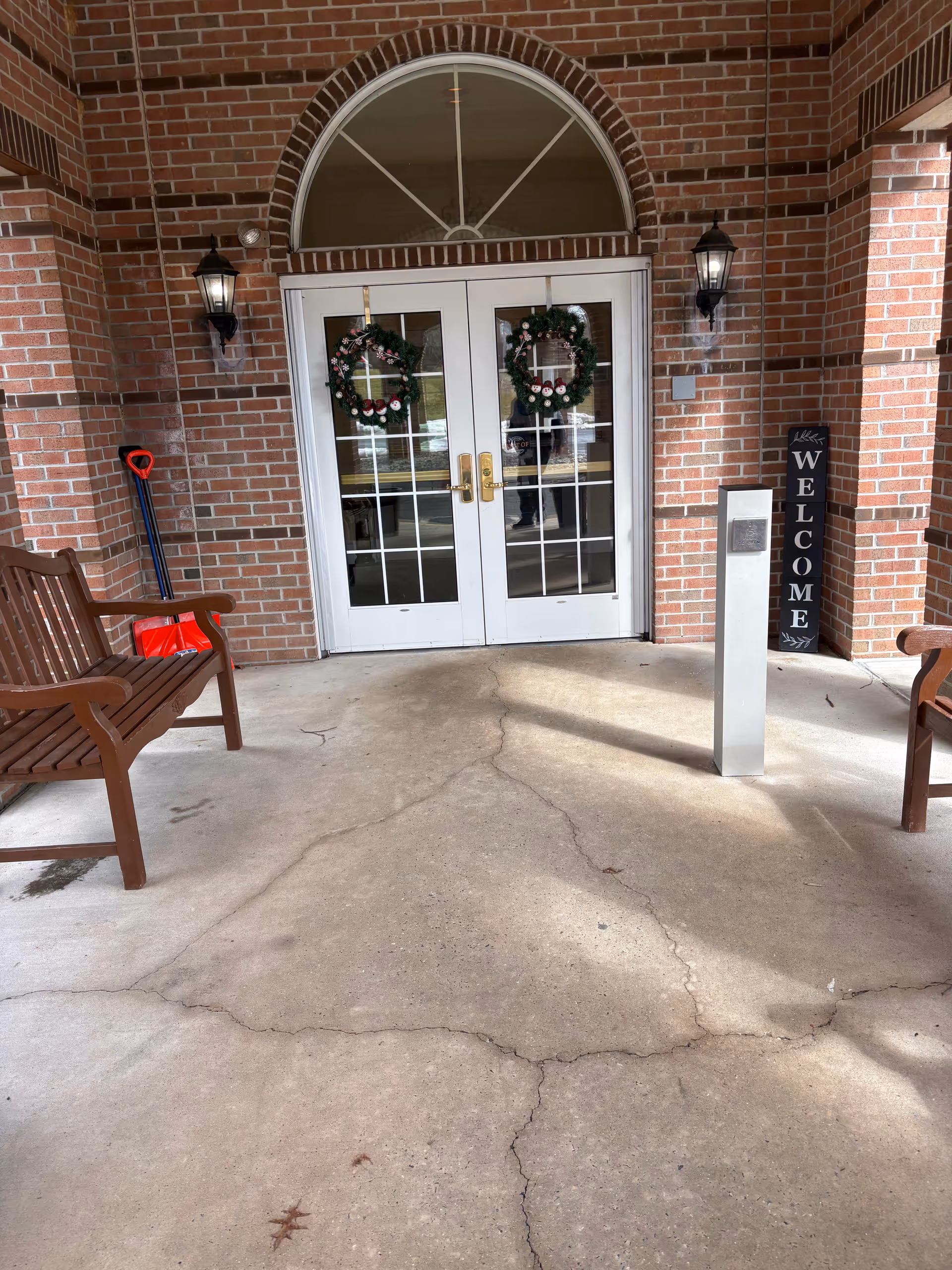 Brick building entrance with double doors, festive wreaths, and wooden benches on a covered porch.