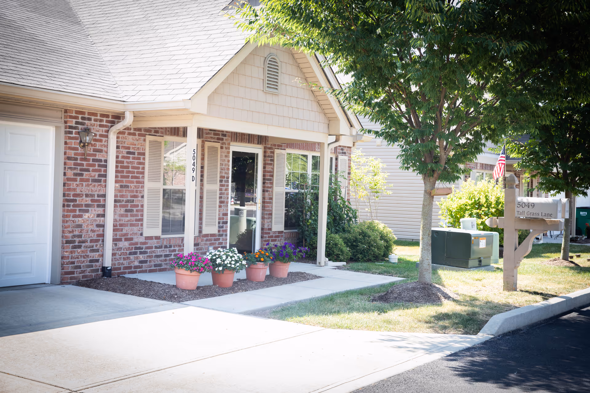 Brick-and-siding assisted living facility entrance with potted flowers and a sign along a neighborhood street.