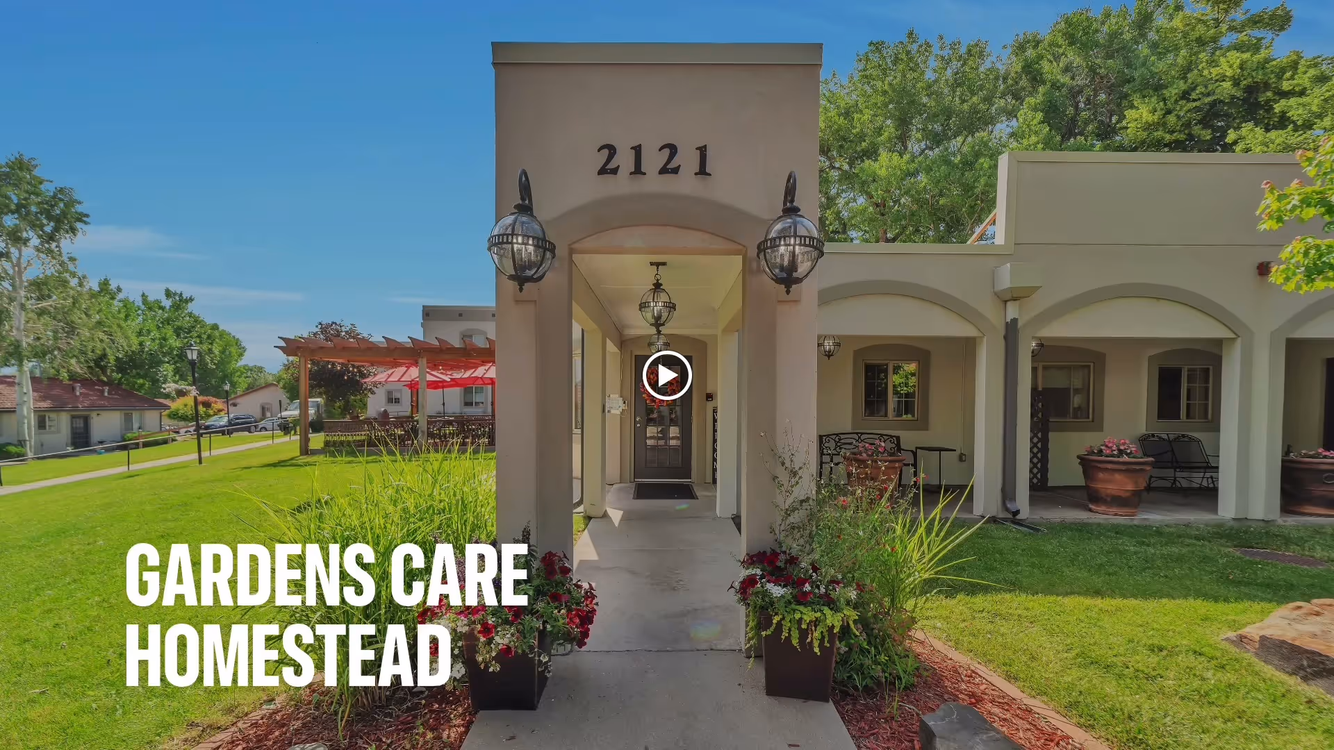Entrance of Gardens Care Senior Living - Homestead facility with a covered walkway, two large lantern-style lights, potted plants with flowers on either side of the path, and a well-maintained lawn and garden area under a clear blue sky.