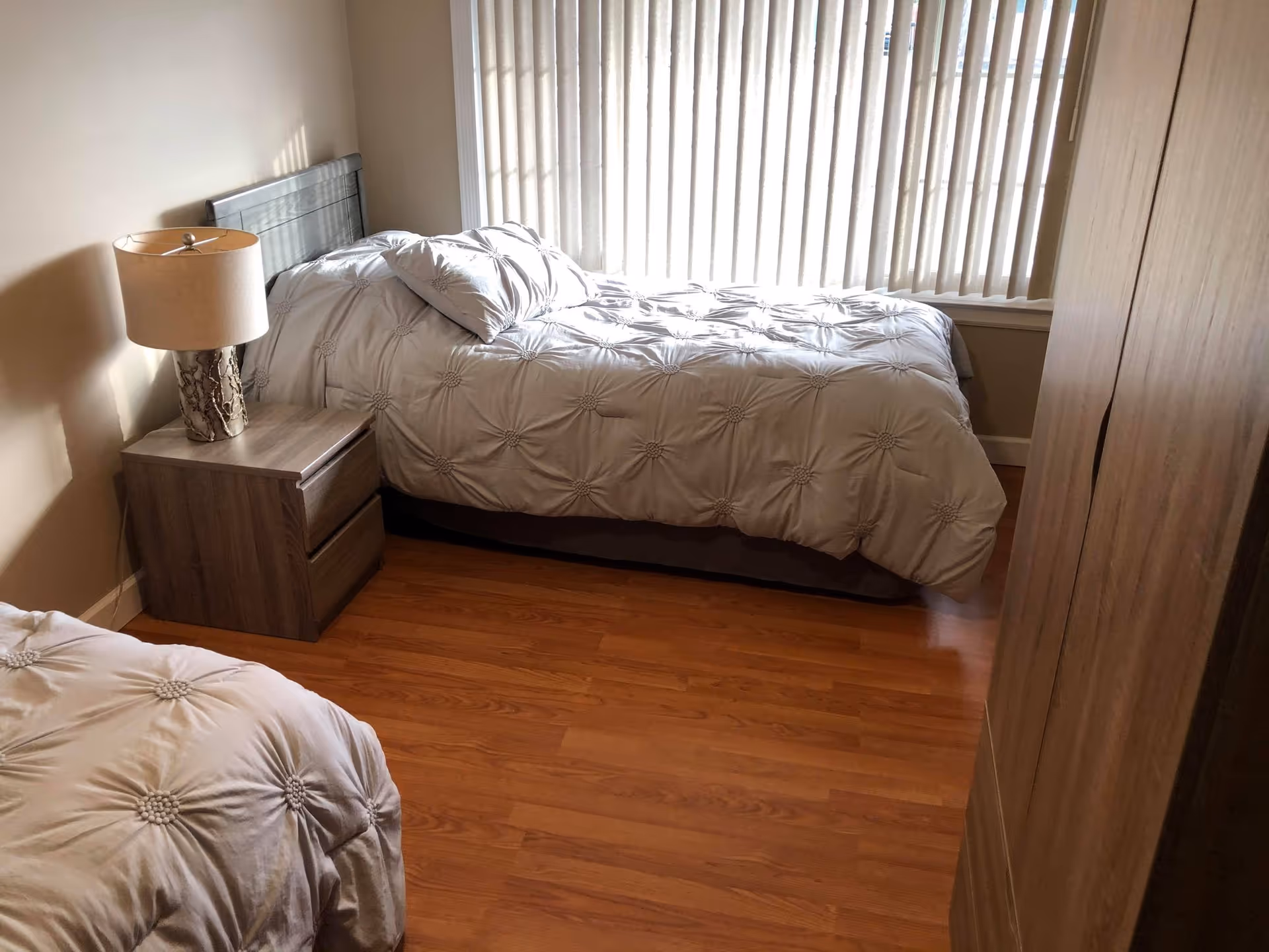 A bedroom with two single beds covered in light gray comforters with a floral pattern. There is a wooden nightstand with a lamp between the beds, a large window with vertical blinds, and a wooden wardrobe on the right side. The floor is wooden.