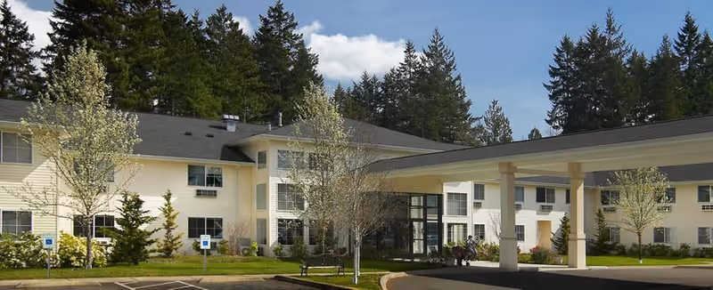 Exterior view of Maple Glen Assisted Living facility showing a two-story building with cream-colored siding, multiple windows, a covered entrance, landscaped greenery, and tall trees in the background under a partly cloudy sky.