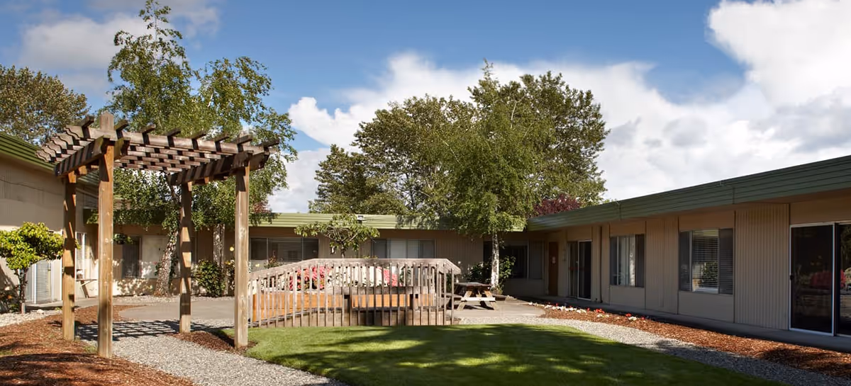 Exterior view of a single-story senior living facility courtyard with a pergola and picnic table.
