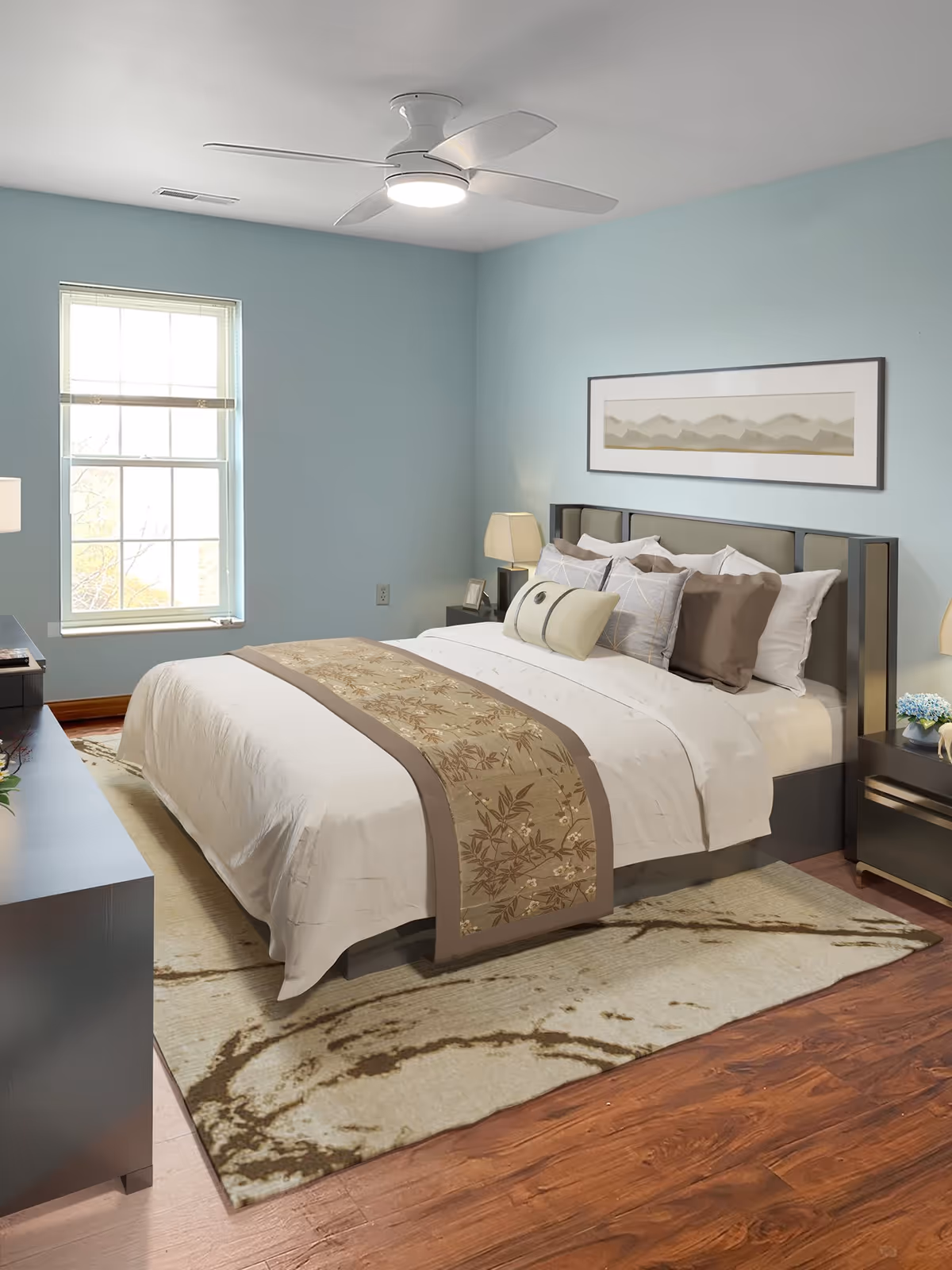 A neatly made bed with white and brown bedding in a bedroom with light blue walls. There is a ceiling fan with a light above the bed, a window with blinds on the left wall, and two nightstands with lamps on either side of the bed. A framed landscape artwork hangs above the headboard. The floor is wooden with a beige and brown patterned rug under the bed.