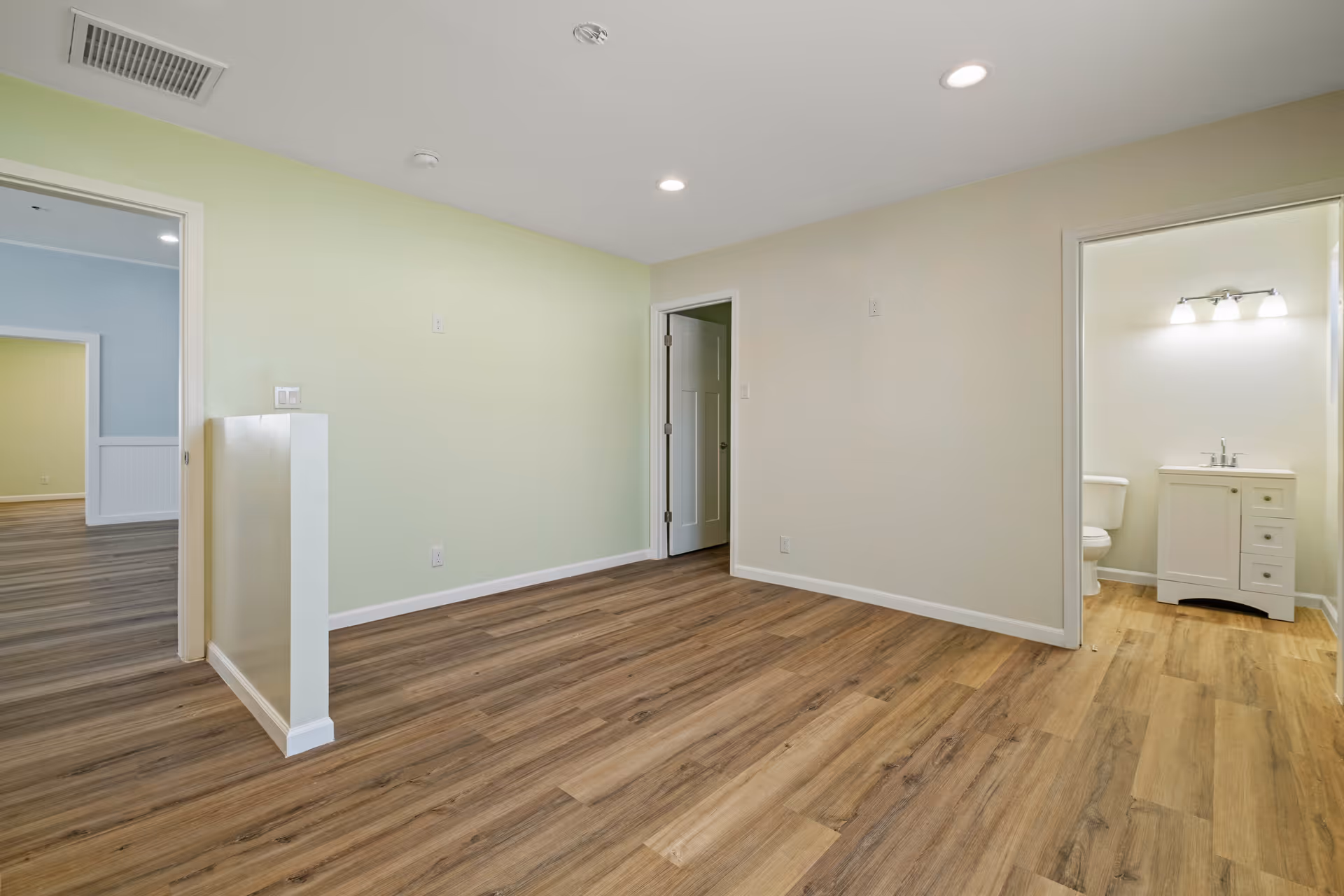 Empty interior room with wood flooring, pale green walls, and doorway to a bathroom.