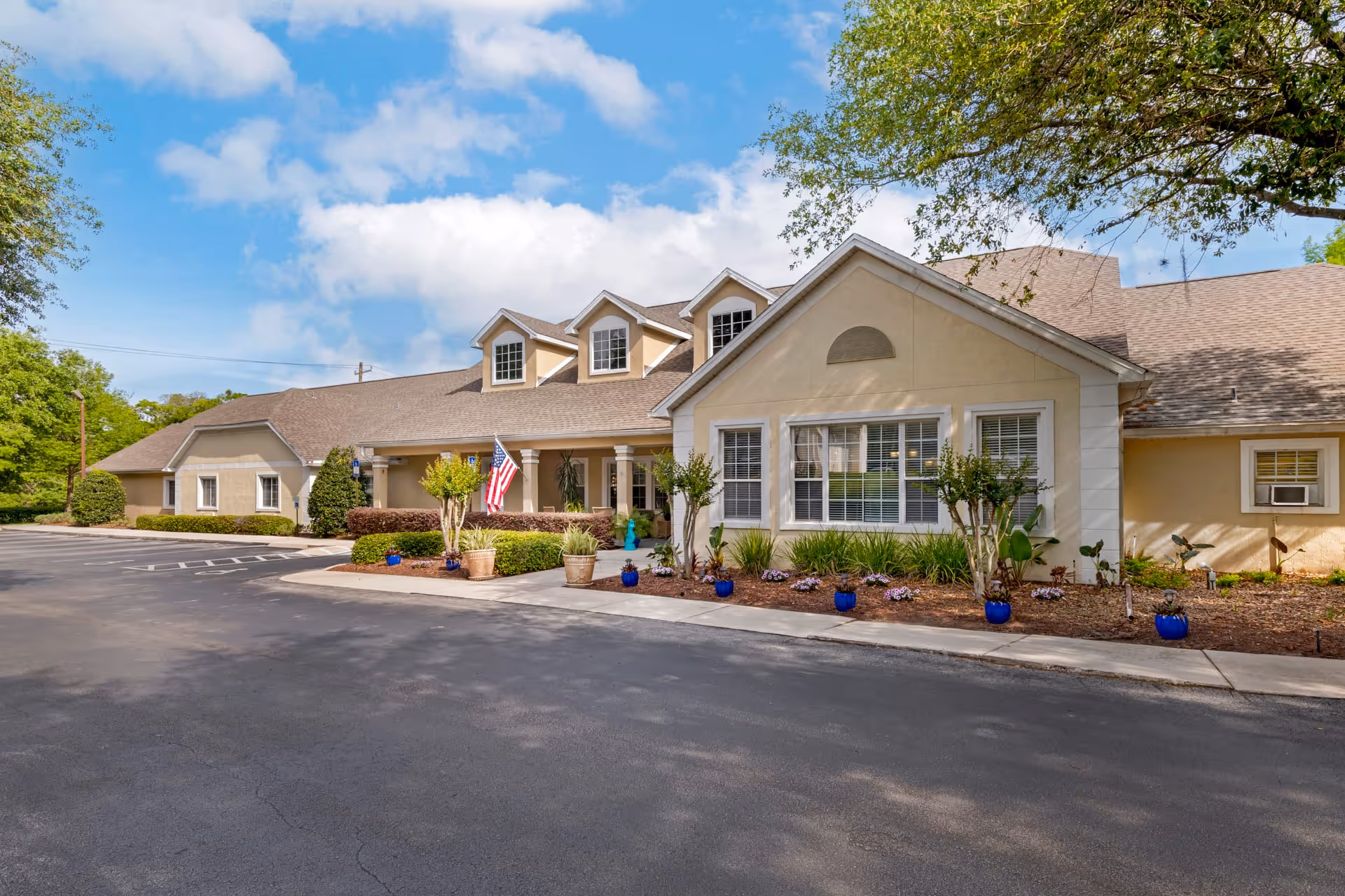 Exterior view of a single-story senior living facility building with beige walls and a brown shingled roof. The building features several windows, a covered entrance with columns, and an American flag displayed near the entrance. There are small trees, shrubs, and potted plants arranged along the front of the building, and a paved driveway and parking area in front. The sky is partly cloudy with blue patches visible.