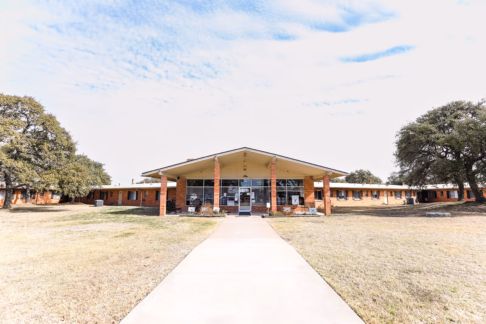 Wide view of a single-story brick senior living facility with a central entrance and lawn.