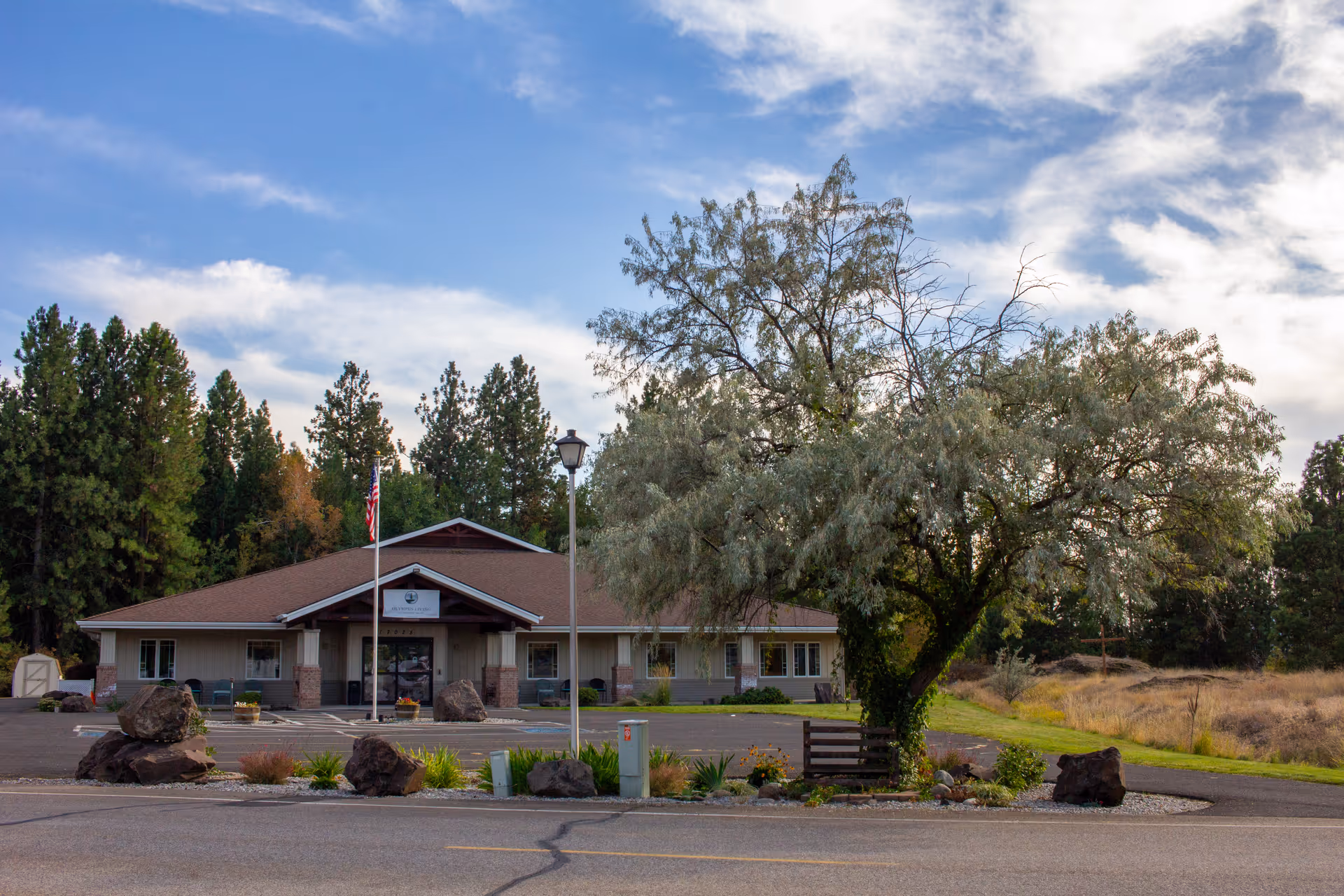 Exterior view of a single-story senior living facility with a flag, trees, and a parking lot.