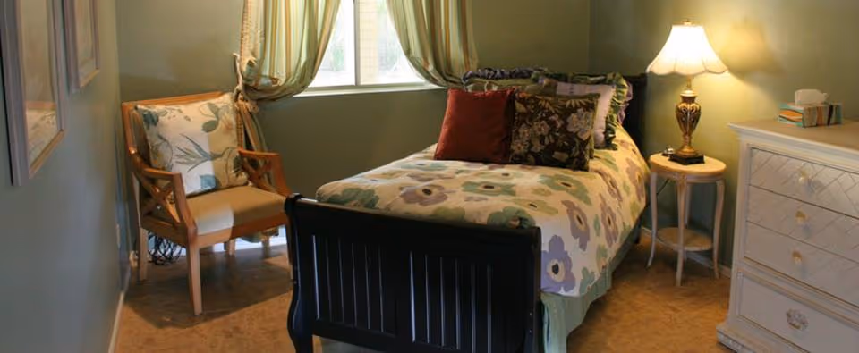 Small bedroom with a single bed dressed in floral bedding, an armchair by a curtained window, a nightstand with a lamp, and a white dresser.