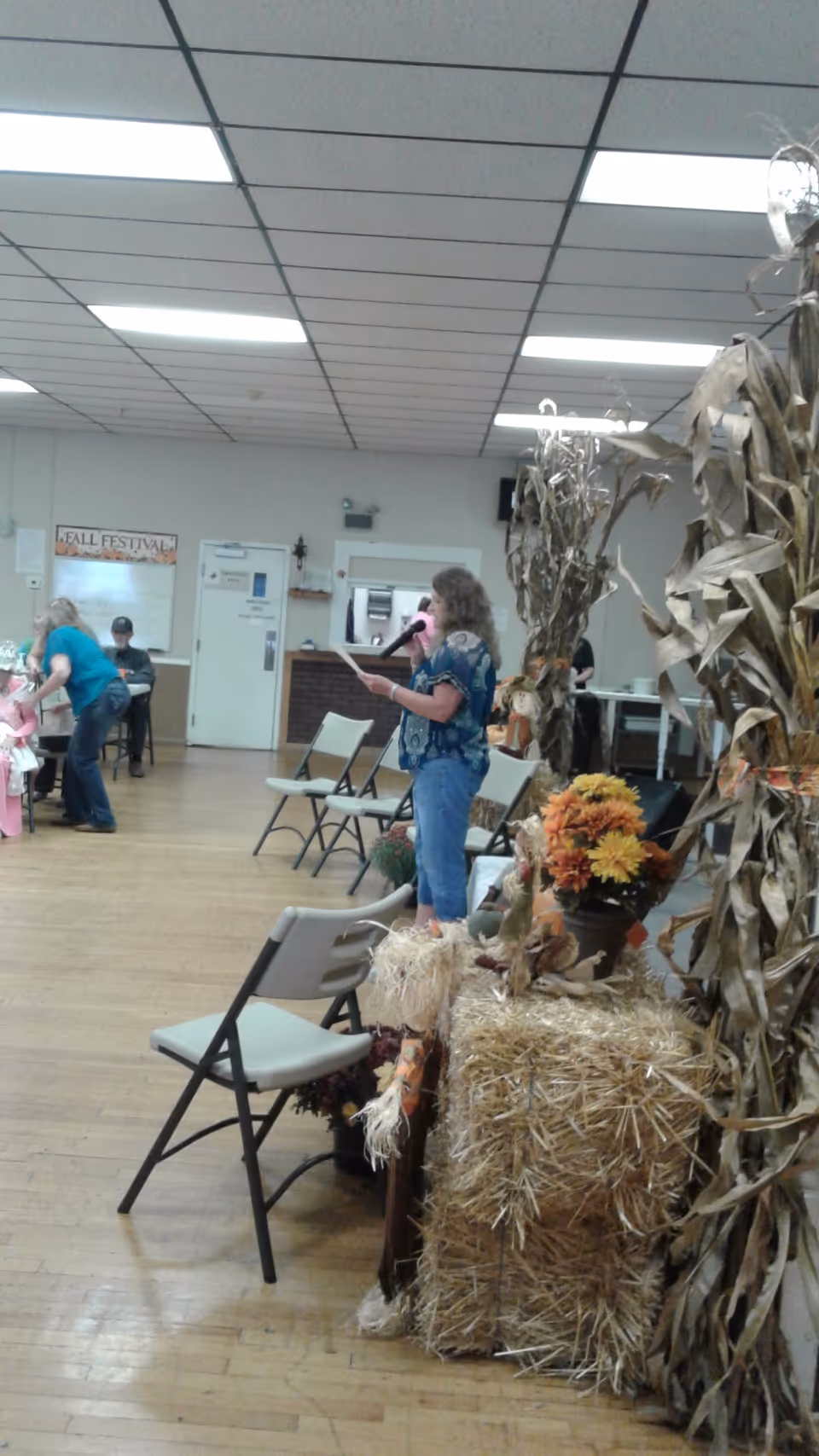 A woman speaks into a microphone in a decorated community hall.