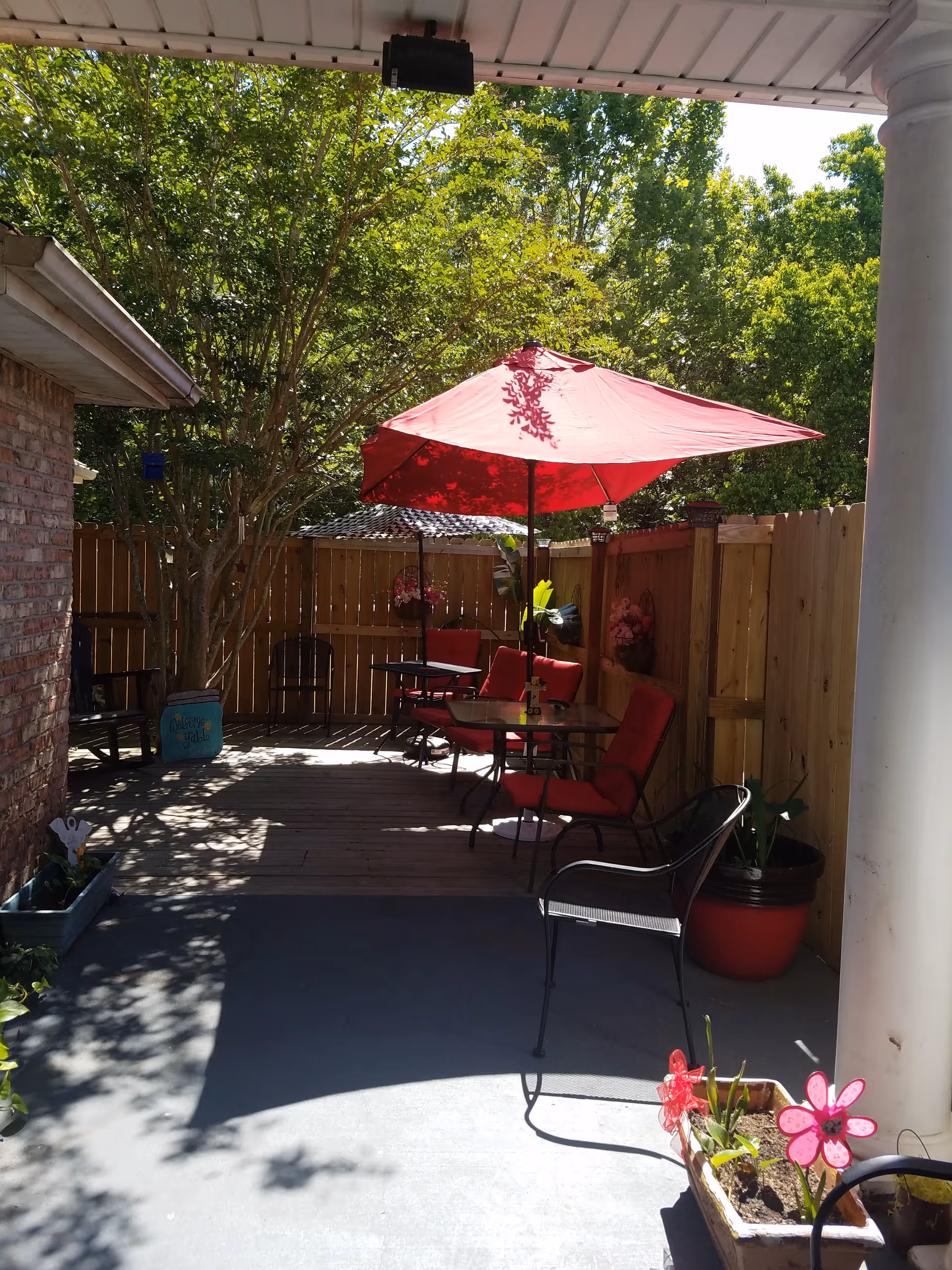 Outdoor patio area with a wooden deck, red cushioned chairs, and a red umbrella providing shade. The space is enclosed by a wooden fence and surrounded by green trees. There are potted plants and decorative items, including a welcome sign and a pink flower decoration in a planter.